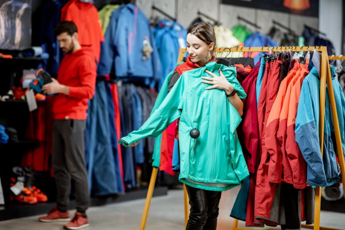 Women trying on jacket at a clothing store while a man looks at shoes behind her.